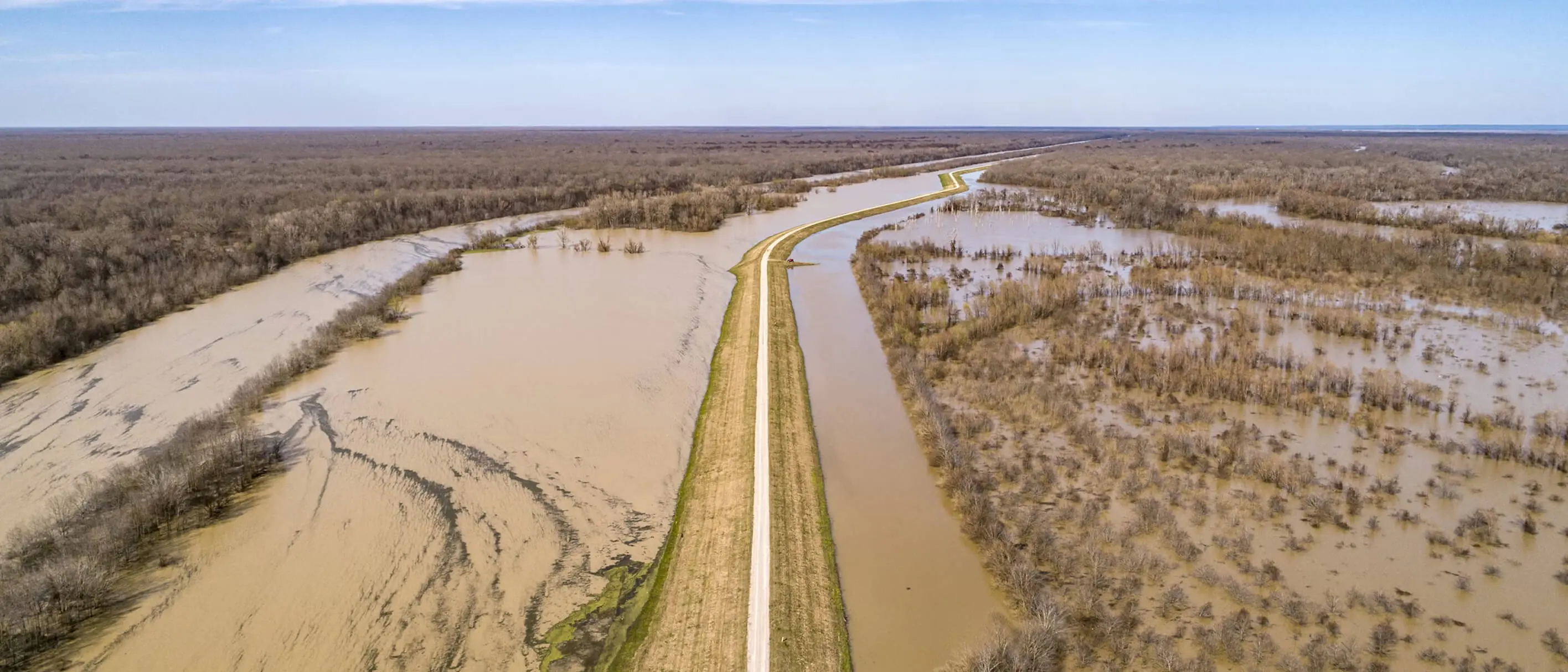 A road on a raised bank cuts through a floodplain cover with brown floodwater with trees sticking out of the flood.